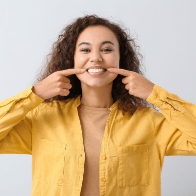 Woman wearing yellow shirt, pointing to her teeth with both hands, representing preventive dental care