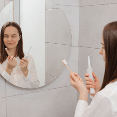 Woman  holding both electric and manual toothbrushes, standing in front of a mirror, representing electric vs manual toothbrush