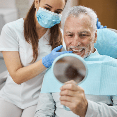 Older man looks in a mirror at his new teeth, while dental professional looks in the background, representing all-on-x implants