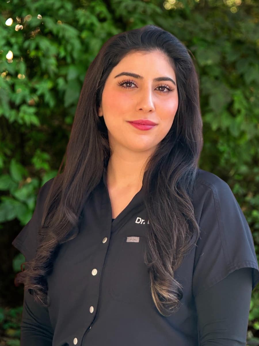 Professional headshot of dr. Elma parsa wearing black scrubs, standing outdoors in front of green foliage.